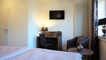 Habitación de hotel con una cama, un escritorio con lámpara, una silla, una mesita y un televisor en la pared.