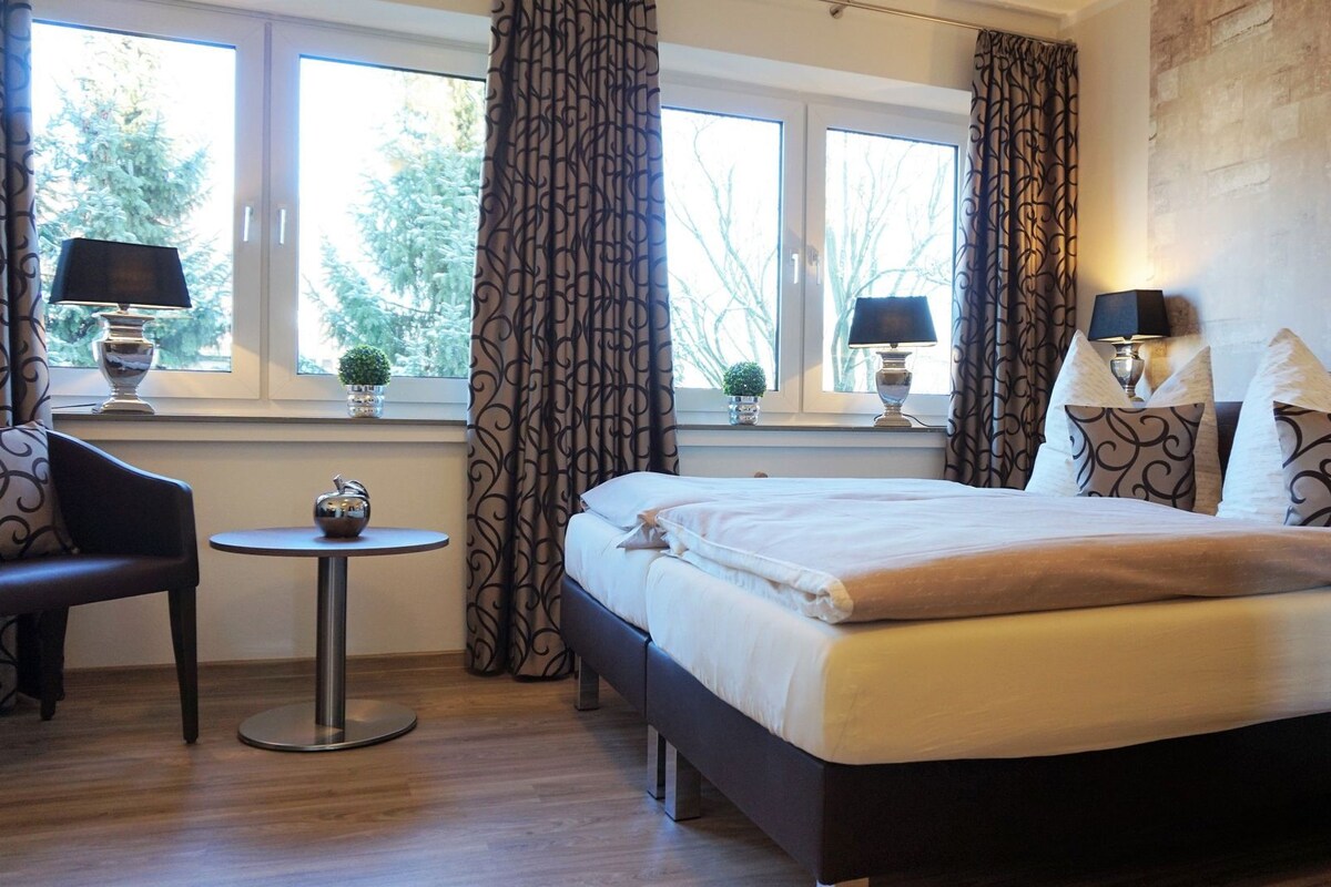 A hotel room with a bed, two bedside lamps, a small round table, a chair, and a view of trees through the window.