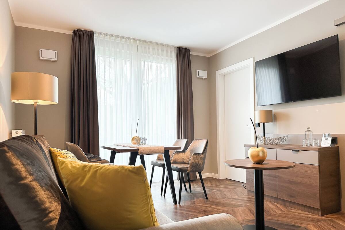 A modern hotel room with a sofa, a dining table, chairs, a TV, and a minibar.