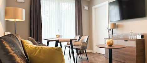 A modern hotel room with a sofa, a dining table, chairs, a TV, and a minibar.