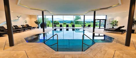 Piscina coberta moderna com vista ampla para o exterior, cercada por espreguiçadeiras e plantas em vasos.