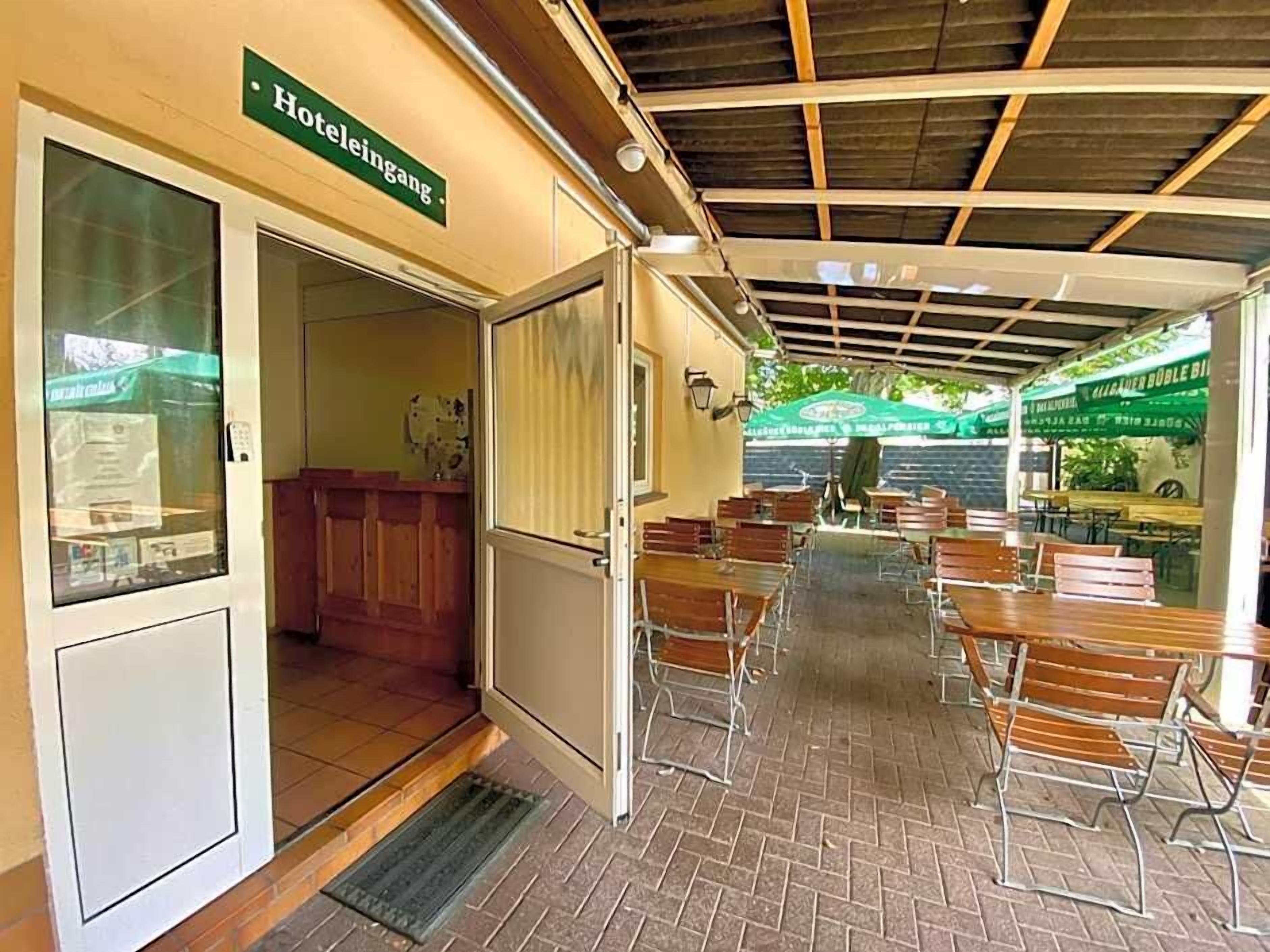 Entrada de hotel con un cartel, área de recepción y una zona de estar exterior cubierta con mesas y sillas.