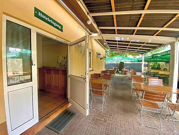 A hotel entrance with a sign, a reception area, and a covered outdoor seating area with tables and chairs.