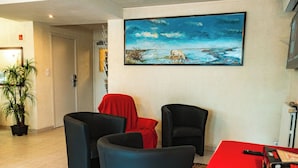 A lobby with a painting of a landscape, a potted plant, and two black chairs with a red tablecloth. - Logis Hotel Heraclee (Saint-Gilles)