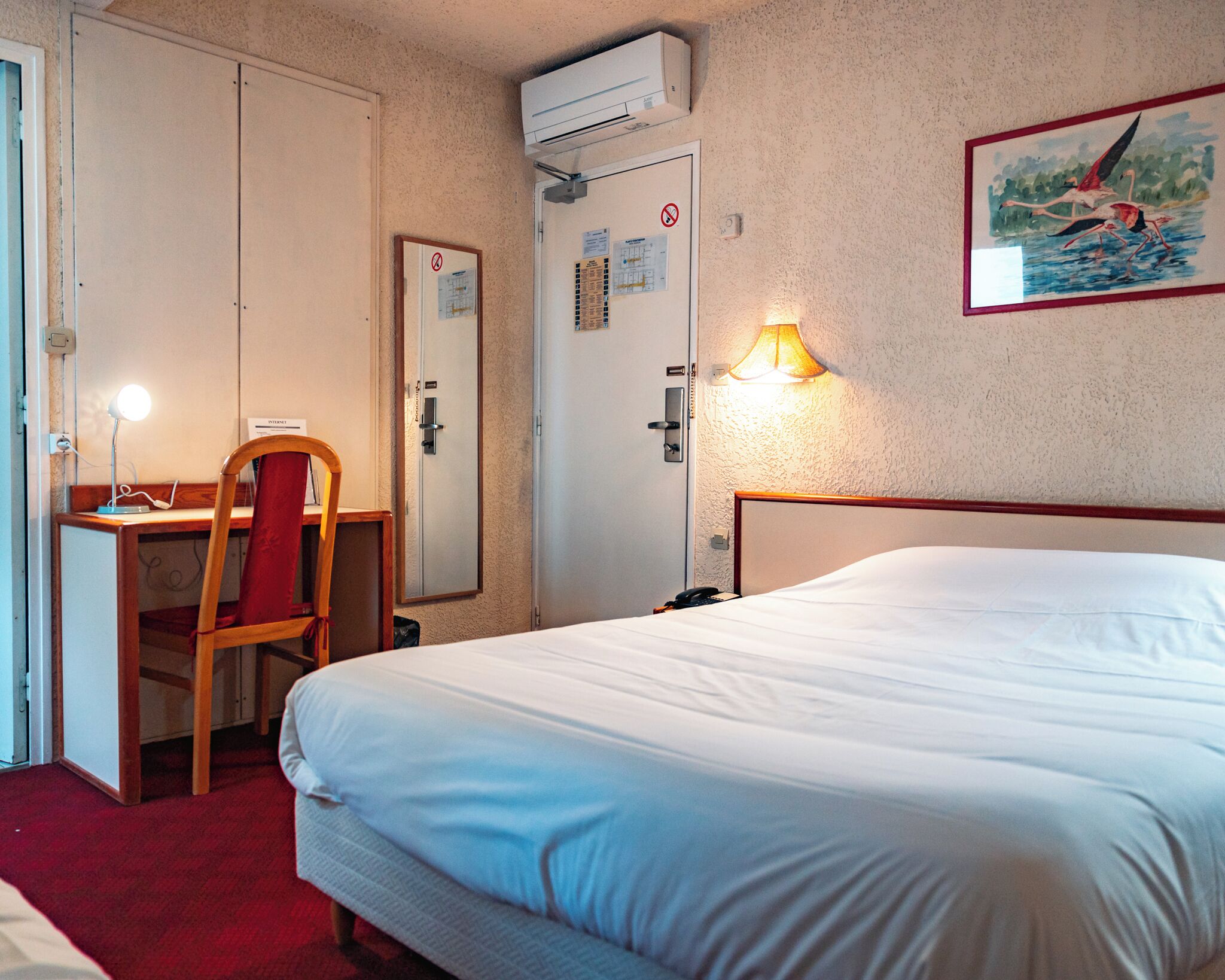 A hotel room with a bed, desk, chair, and a painting on the wall.