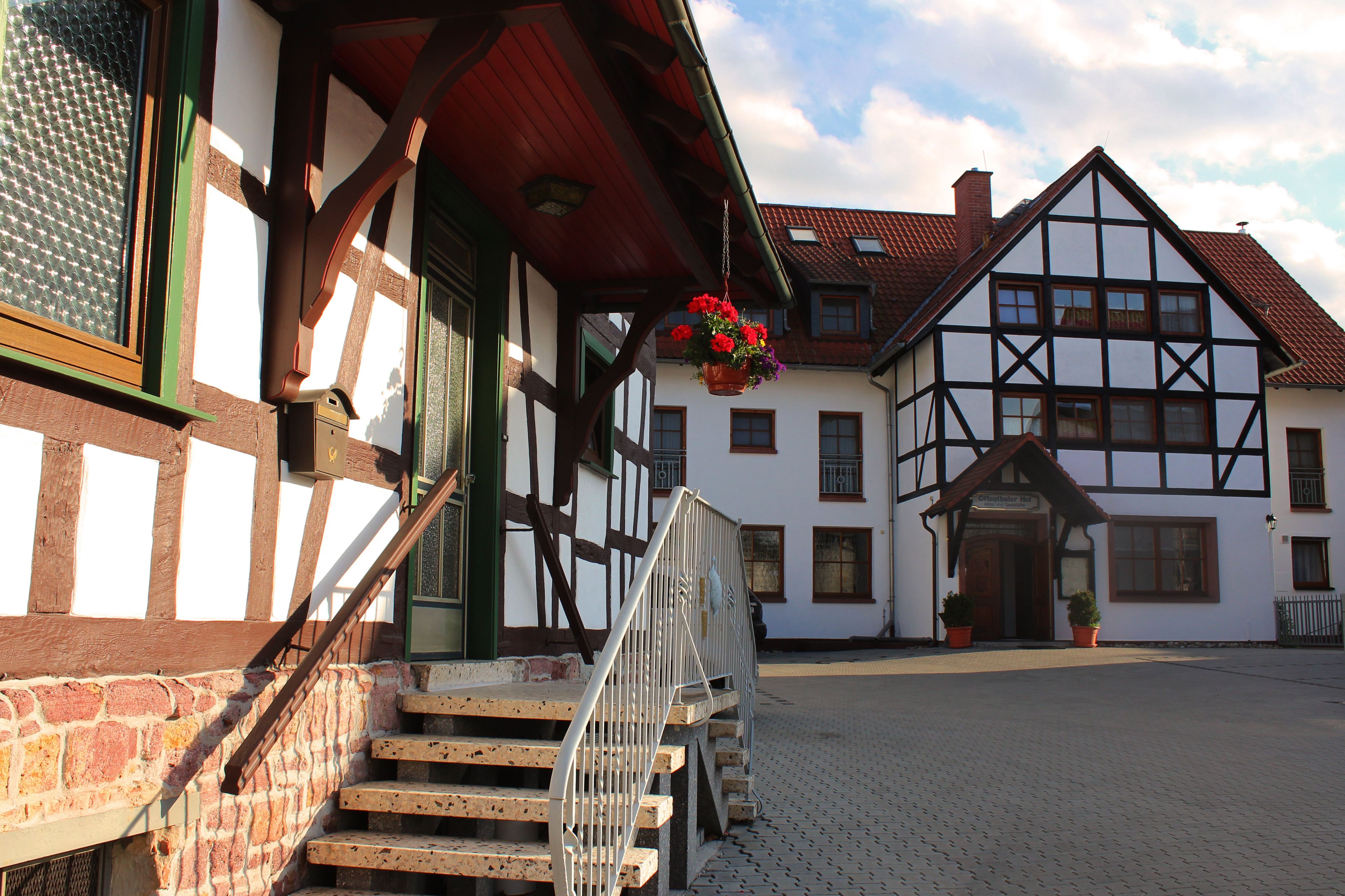 A traditional half-timbered building with a staircase leading to a wooden door, a hanging flower basket, and a paved courtyard.