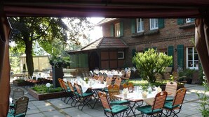 Eine Terrassengaststätte mit Tischen und Stühlen, im Hintergrund ein Backsteinhaus und Grünpflanzen.