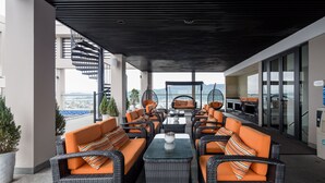 Un espacio al aire libre con mobiliario de mimbre y cojines anaranjados, con vista a un paisaje urbano.