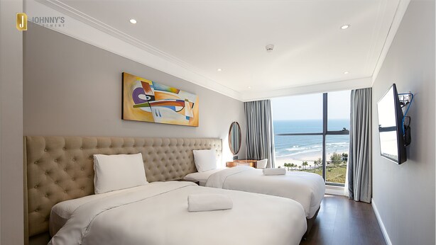 Habitación de hotel con dos camas, un ventanal con vista al océano, un cuadro en la pared y un televisor de pantalla plana montado en la pared.