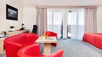 A hotel room with a bed, a desk with a computer, a chair, a table with breakfast, and a balcony with curtains.