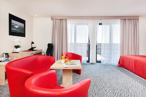 A hotel room with a bed, a desk with a computer, a chair, a table with breakfast, and a balcony with curtains.