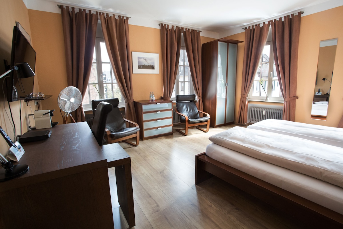 A hotel room with a bed, desk, chairs, a desk lamp, a fan, and a window with curtains.
