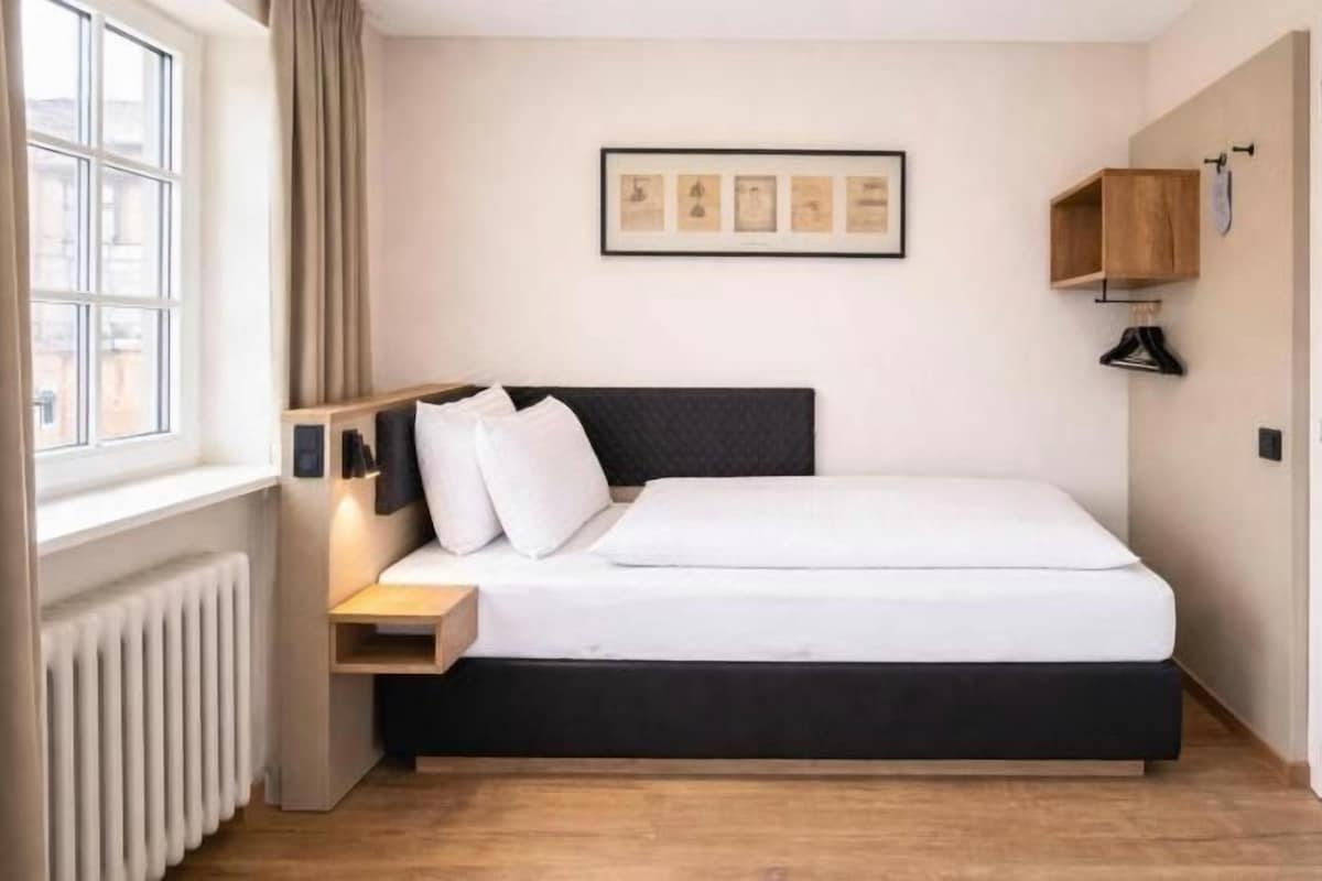 A hotel room with a bed, bedside table, lamp, and a window.