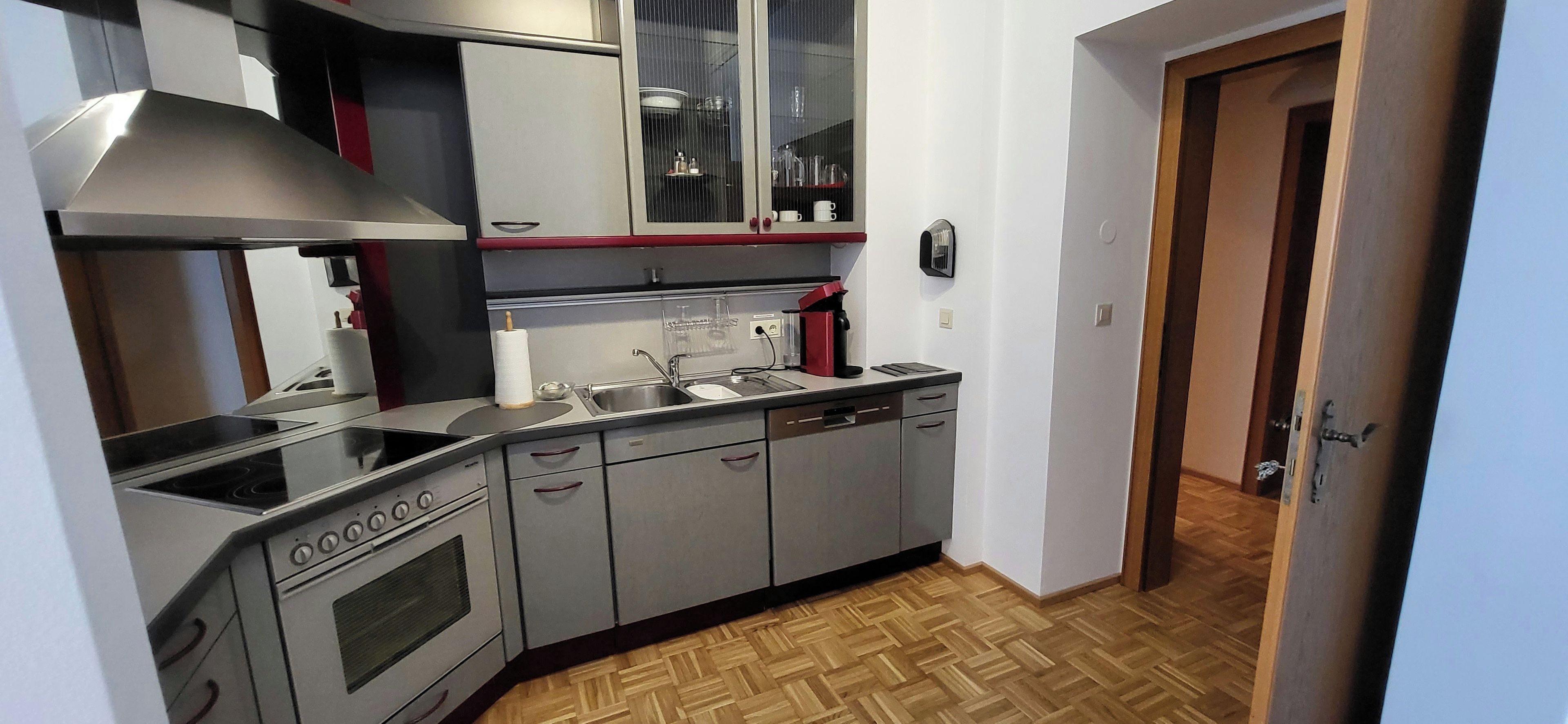 Cocina compacta con estufa, lavamanos y armarios, piso de madera y una puerta que da a otra habitación.