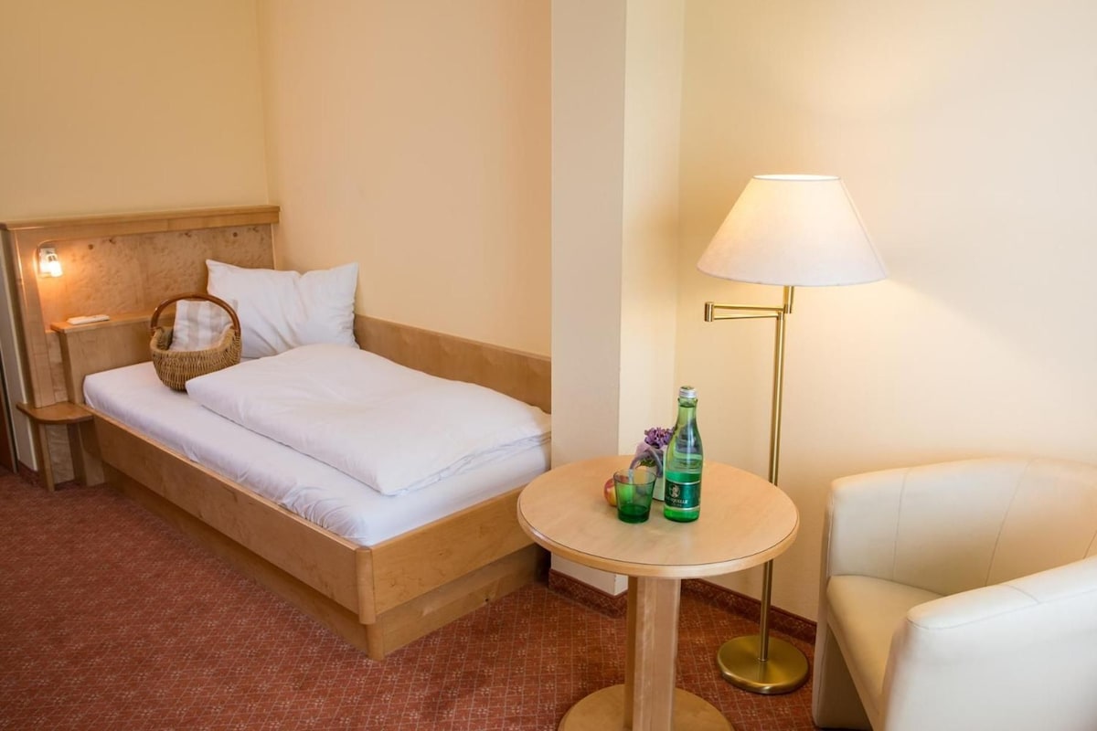 A hotel room with a bed, a bedside table with a lamp, a small round table with bottles, and a chair.
