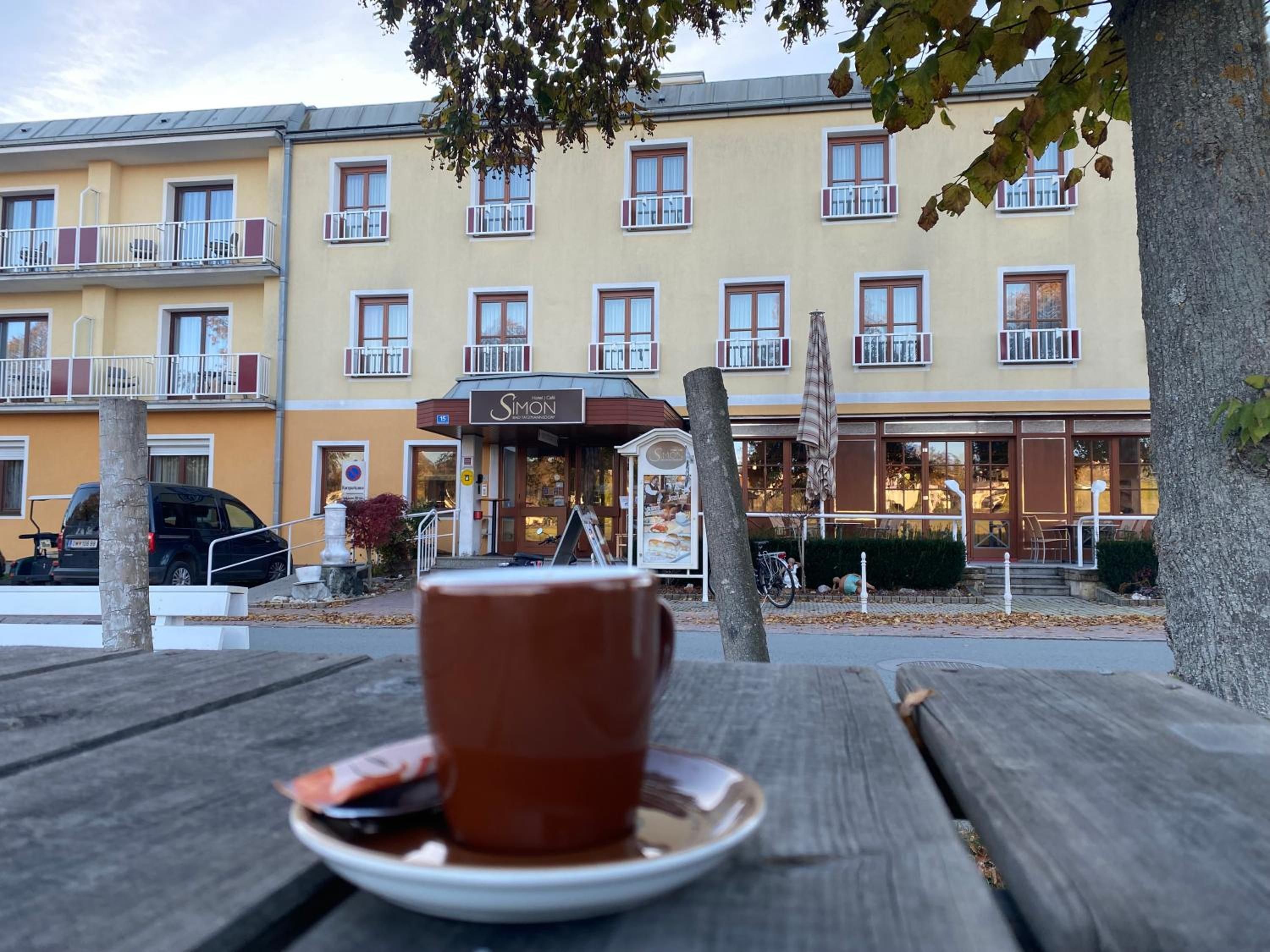 Ein Esstisch mit Tasse und Untertasse, eine Bank und im Hintergrund ein Gebäude mit zahlreichen Fenstern und Balkonen.