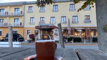 Ein Esstisch mit Tasse und Untertasse, eine Bank und im Hintergrund ein Gebäude mit zahlreichen Fenstern und Balkonen.