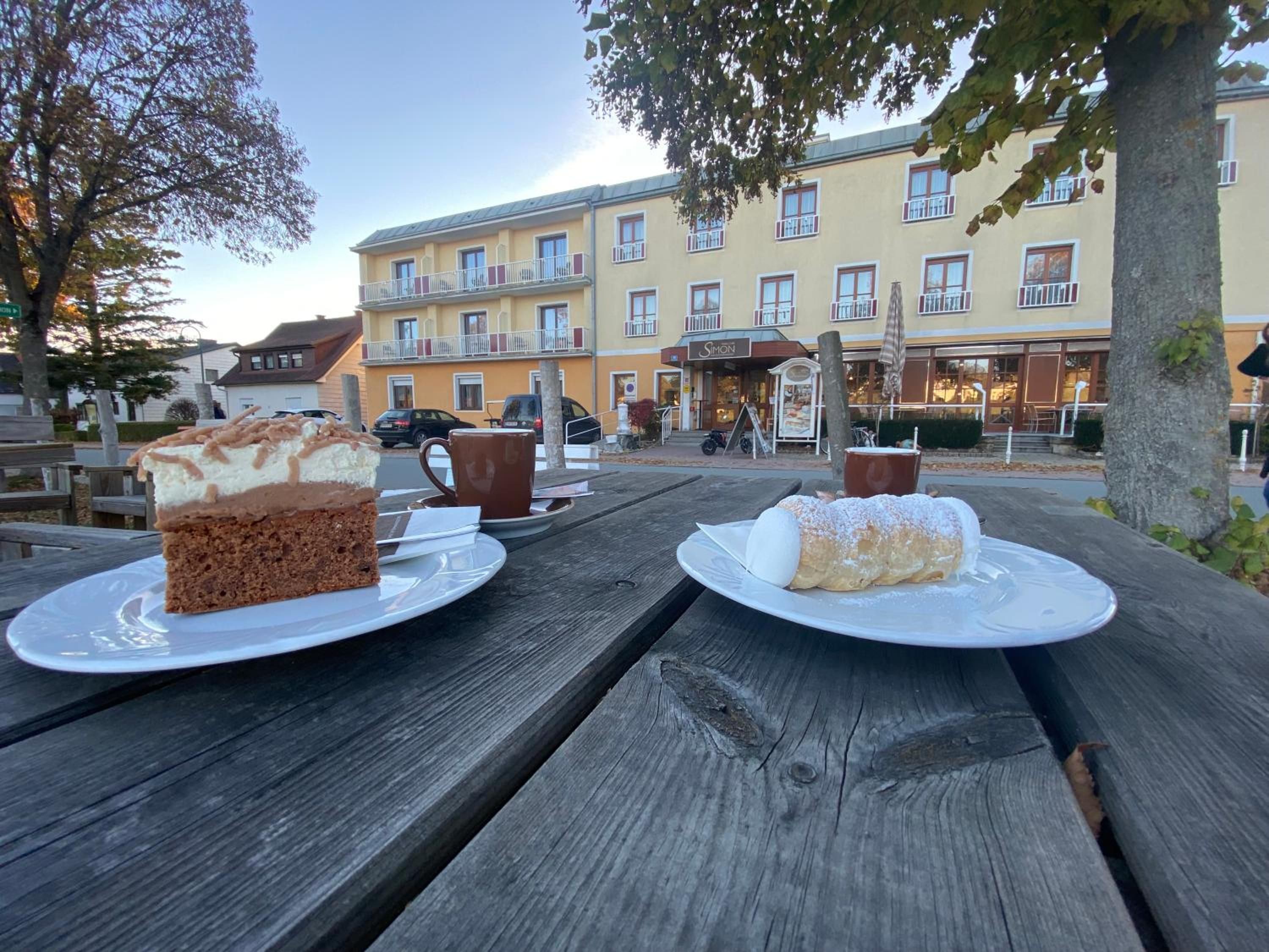 Ein Holztisch für ein Picknick mit zwei Desserttellern, einer Kaffeetasse und einem Schild für das Restaurant 'Simon' im Hintergrund.