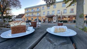 Ein Holztisch für ein Picknick mit zwei Desserttellern, einer Kaffeetasse und einem Schild für das Restaurant 'Simon' im Hintergrund.