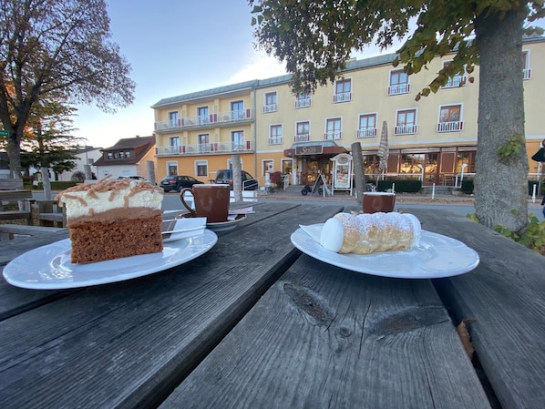 A wooden picnic table with two plates of dessert, a coffee cup, and a sign for a restaurant named 'Simon' in the background. - Simon Hotel Cafv (Bad Tatzmannsdorf)