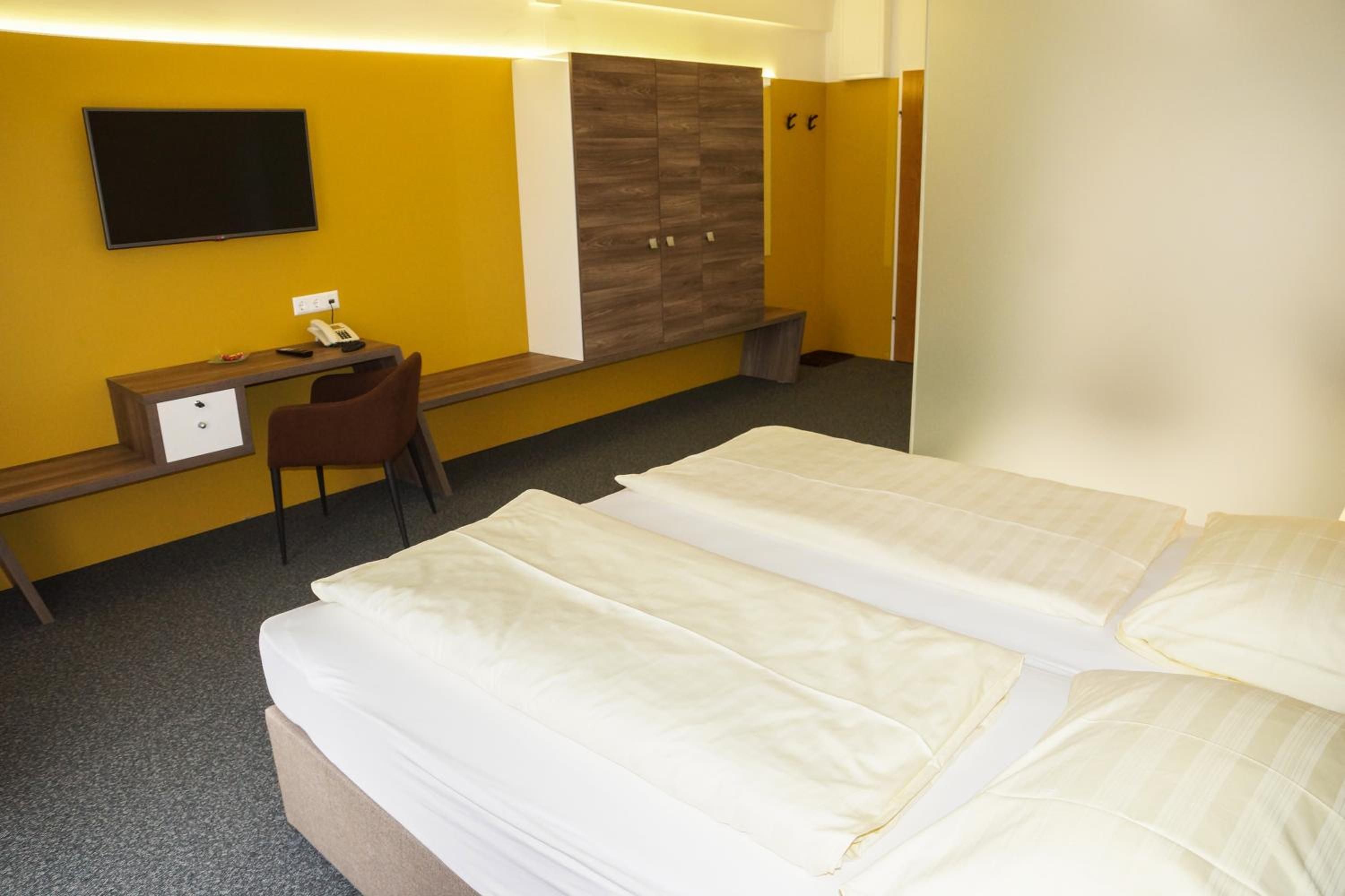 Habitación de hotel con dos camas, un escritorio con silla, un armario y un televisor de pantalla plana montado en la pared.