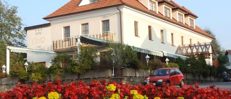 A hotel with a balcony, a red car parked in front, and a flower bed with red and yellow flowers.