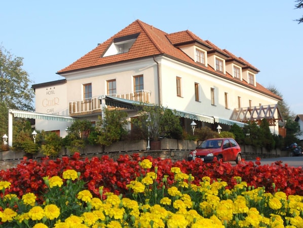 A hotel with a balcony, a red car parked in front, and a flower bed with red and yellow flowers.