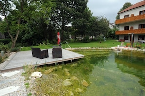 A wooden deck with outdoor furniture, a pond, and a building with a balcony.