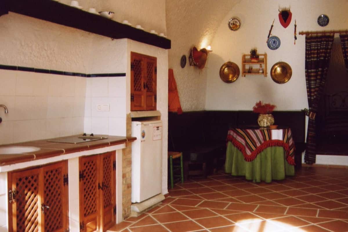Een keuken met een spoelbak, kasten en een koelkast. Er is een eethoek met een tafel en stoelen.