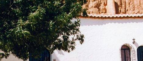 Un edificio blanco con fachada rústica, un árbol en primer plano y una silla verde afuera.