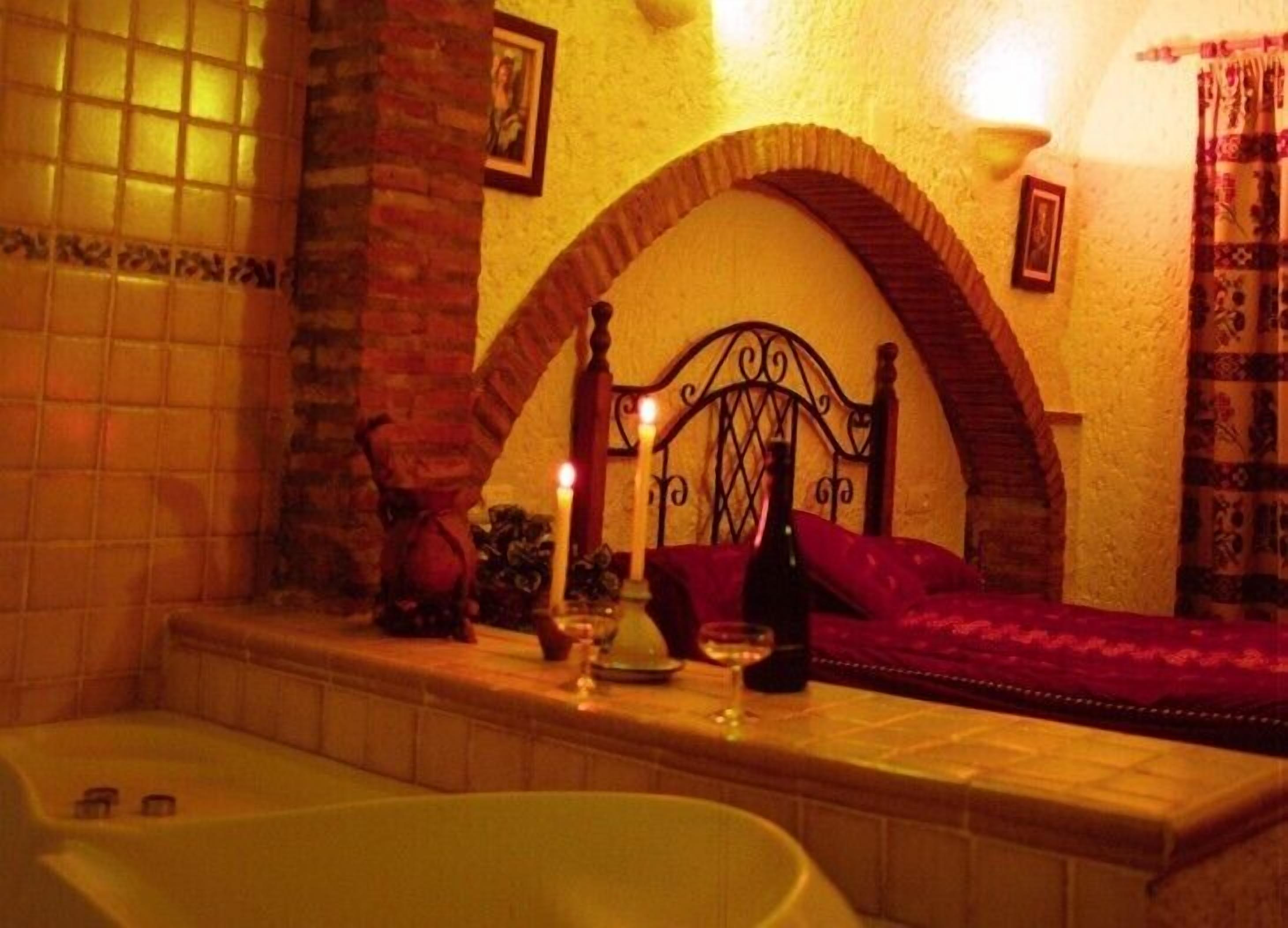 Une pièce avec une baignoire, un lit recouvert d’une couverture rouge, une arche décorative au mur et un tableau accroché au mur.