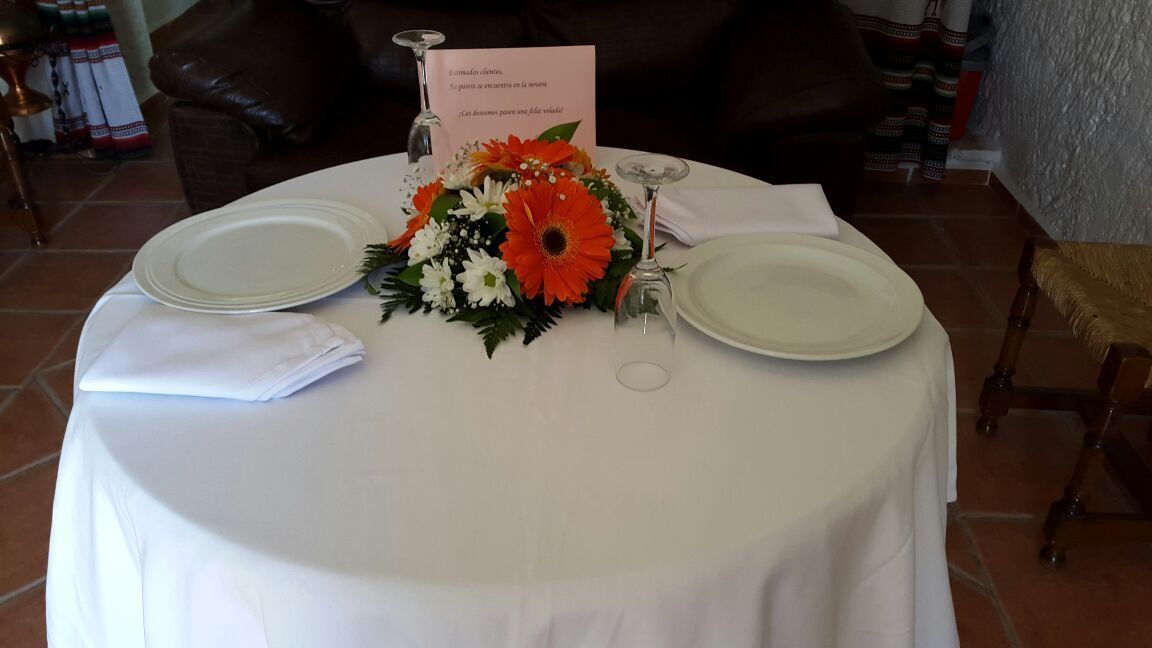 Une table de salle à manger ronde, avec deux assiettes blanches, un centre de table composé de fleurs orange et blanches, et un menu.