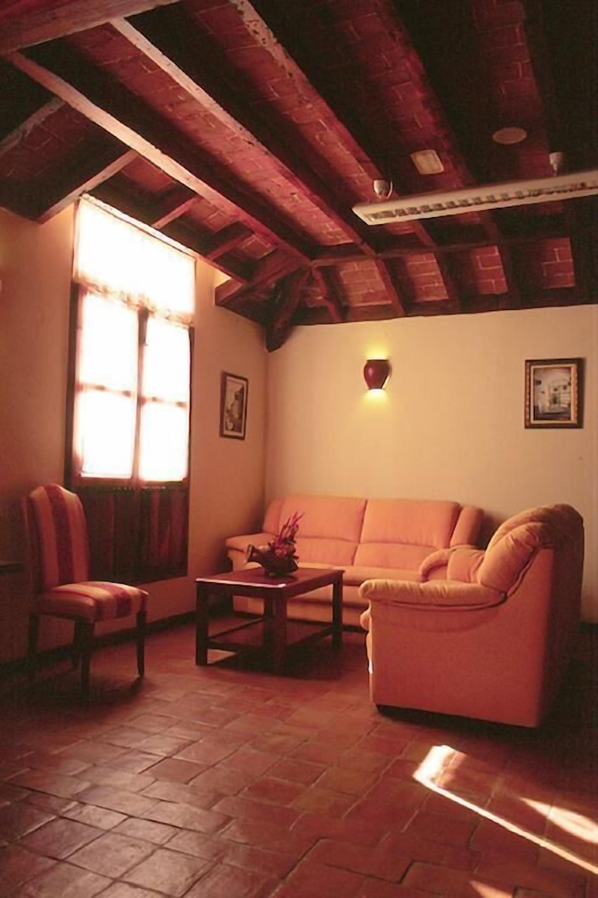 Ein gemütliches Wohnzimmer mit einer Couch, Sesseln und einem Tisch. Es gibt ein Fenster mit Vorhängen, eine Wandleuchte und ein gerahmtes Bild.