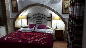 Een bed met een wit hoofdbord, twee kussens en een rode sprei met rozenblaadjes. Twee nachtlampjes en een houten nachtkastje zijn aanwezig.