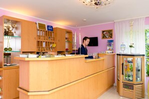 A reception desk in a room with a pink wall, wooden cabinets, and a television mounted on the wall. - 1a Hotel Steiner (Judenburg)