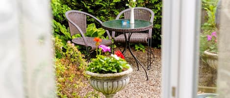 Un espacio al aire libre con sillas de mimbre y una mesa de vidrio, rodeado de follaje y un jarro decorativo.