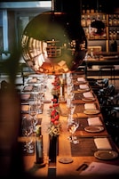Una larga mesa de comedor dispuesta con copas de vino, platos y cubiertos, reflejando un espacio interior bien iluminado.