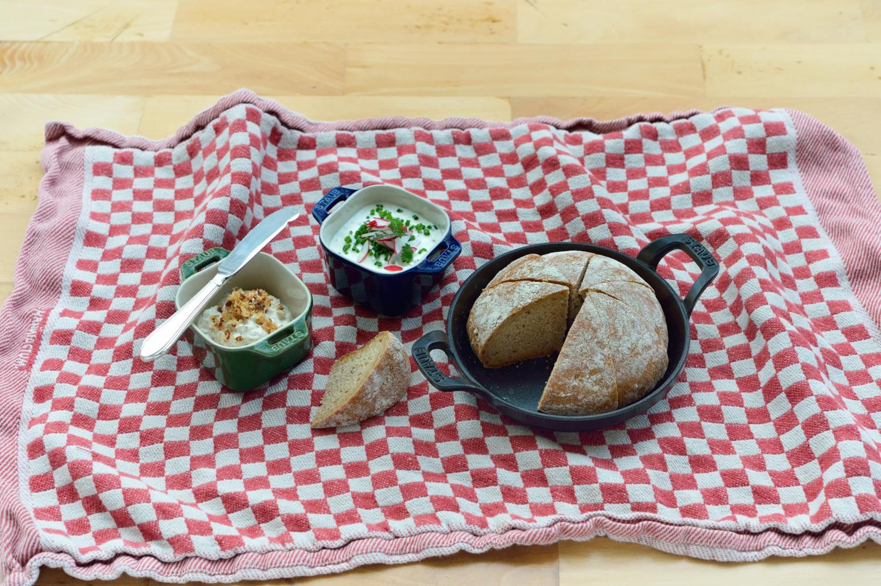 Un mantel a cuadros con una hogaza de pan, un tazón de sopa y un cuenco pequeño de yogur sobre una superficie de madera.