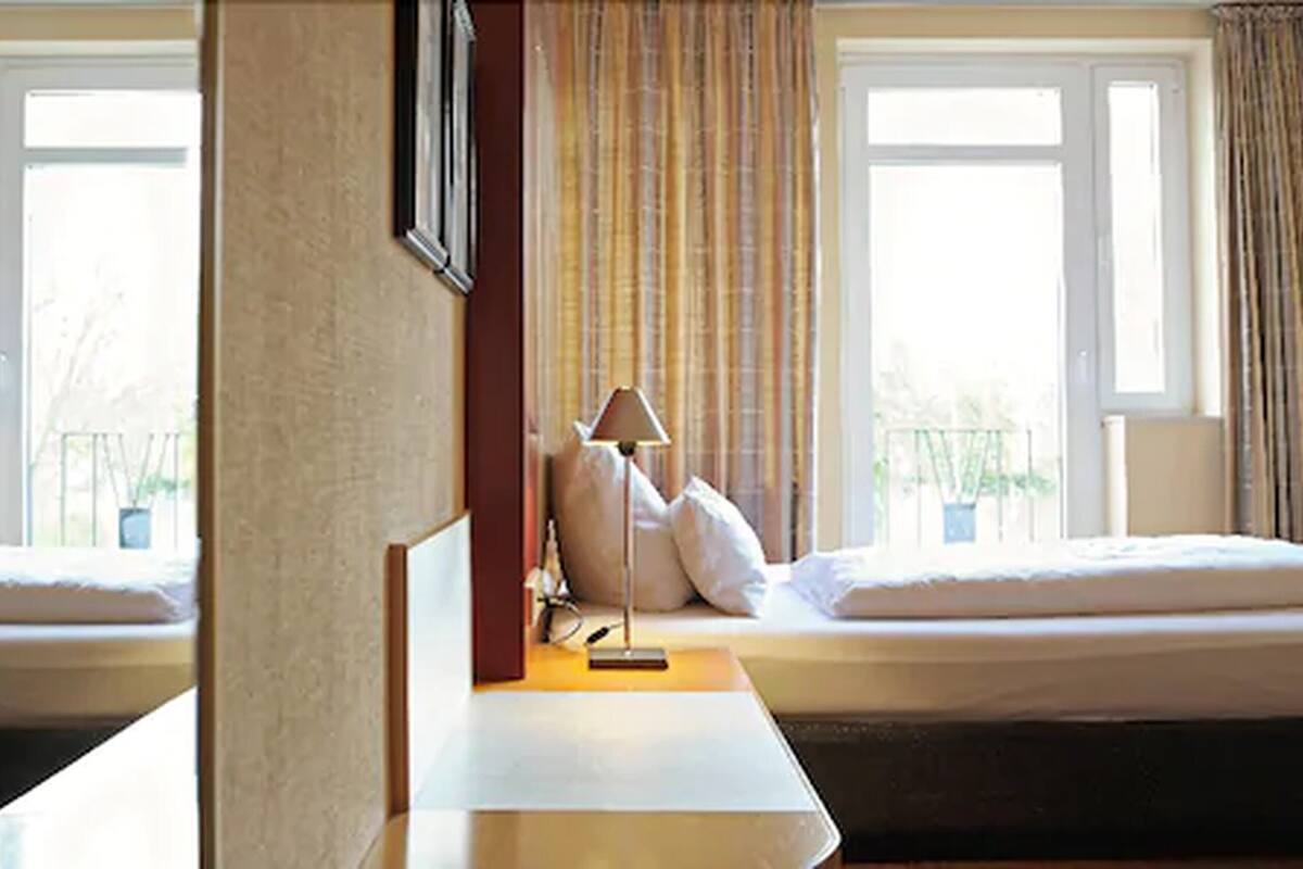 A hotel room with two beds, a bedside table, a lamp, and a window with curtains.