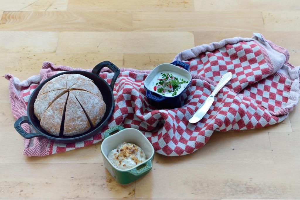 A rustic setting with a loaf of bread, a bowl of soup, and a small dish of what appears to be a creamy dessert, all placed on a checkered cloth.