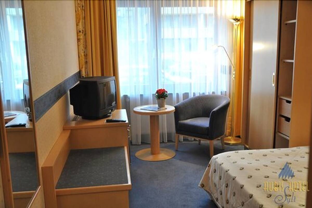 A hotel room with a bed, a desk with a computer, a chair, a small table with a plant, and a TV.