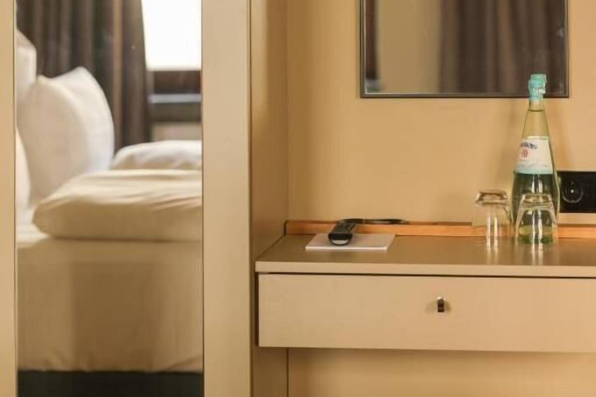 A hotel room with a bed, a bedside table with a bottle, a remote control, and a mirror on the wall.