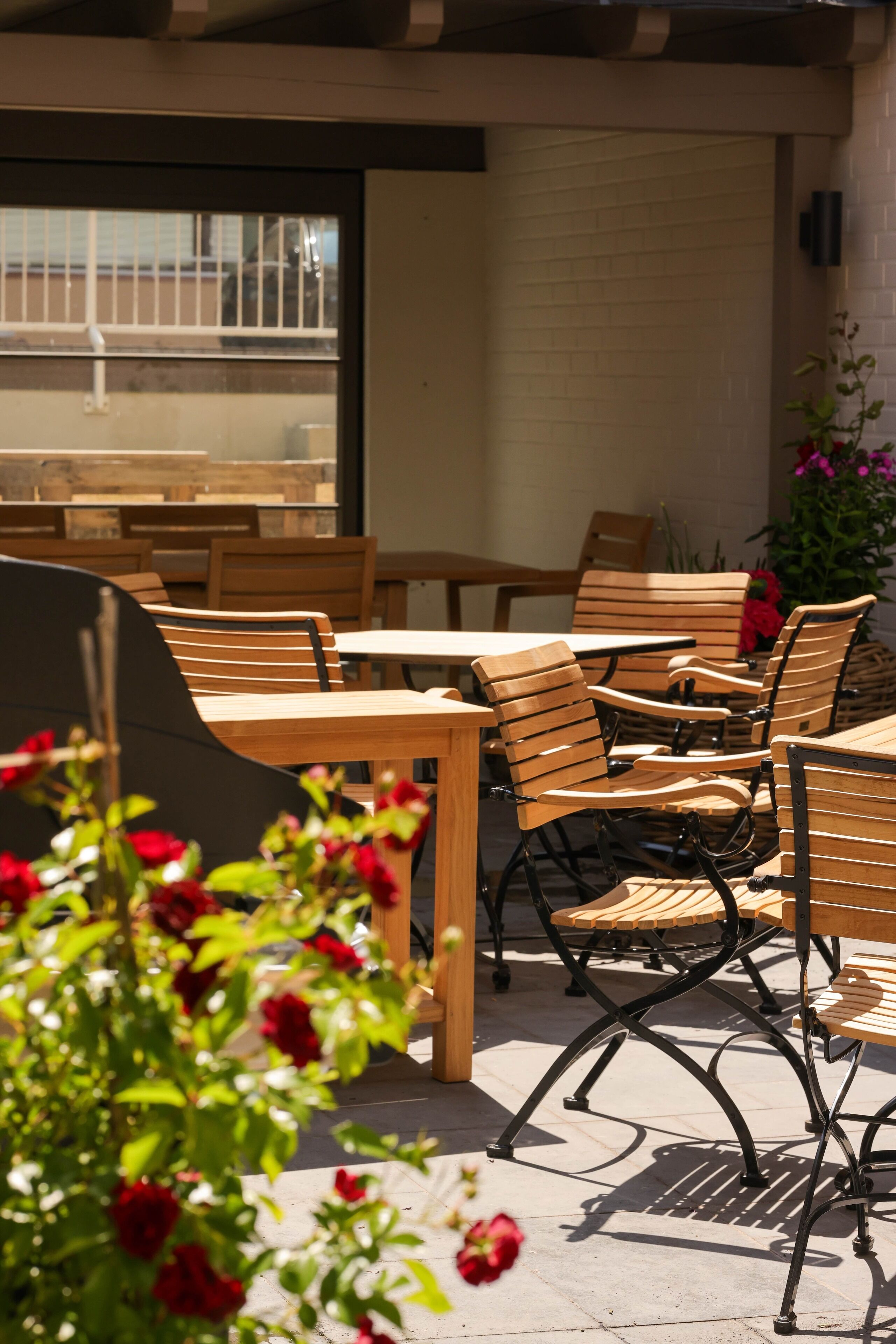 An outdoor seating area with wooden tables and chairs, a brick wall, and a window with a view of a building.