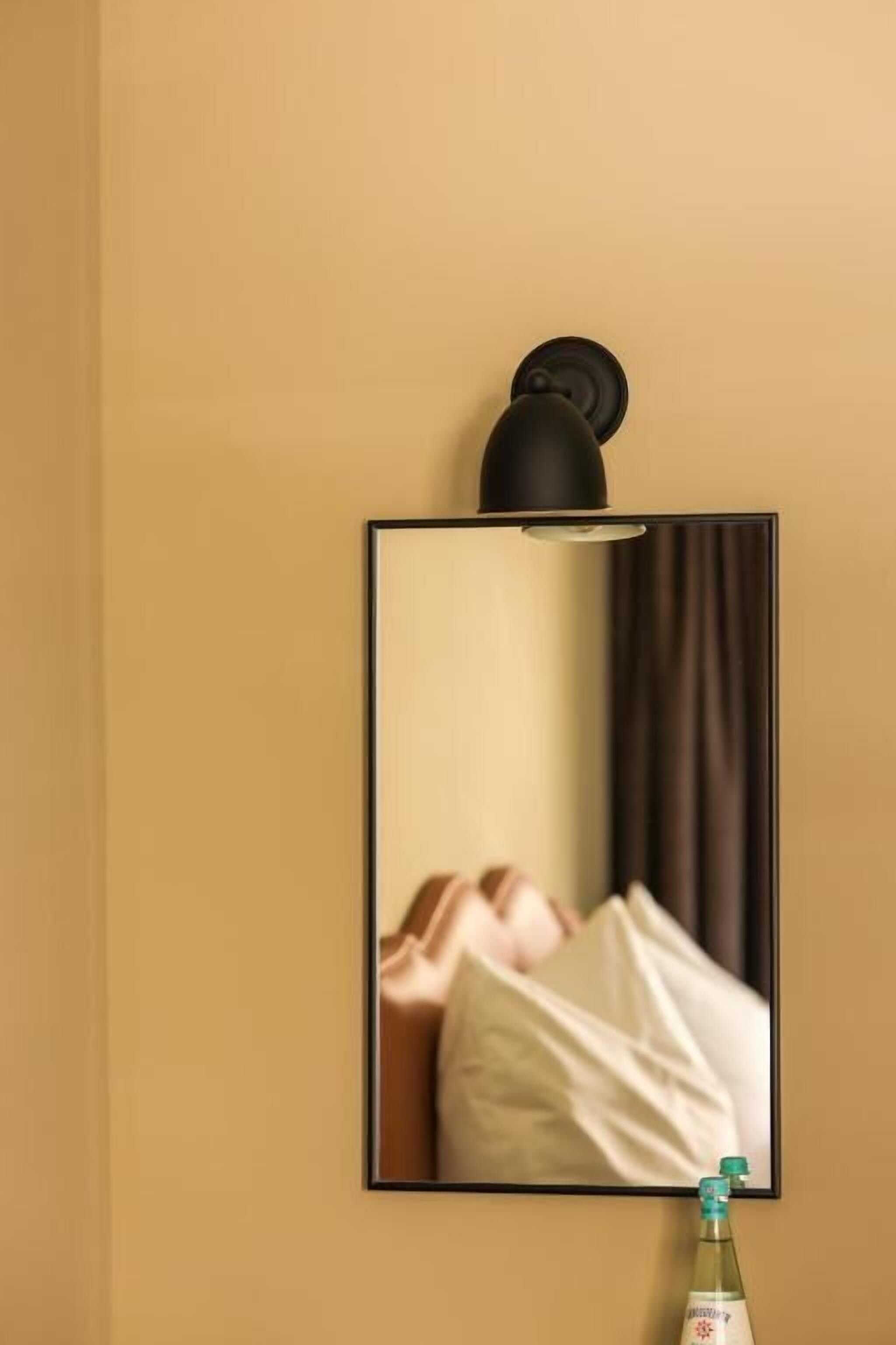 Fotografía enmarco de una cama con almohadas, una lámpara fijada a la pared y una botella de agua sobre una mesa.