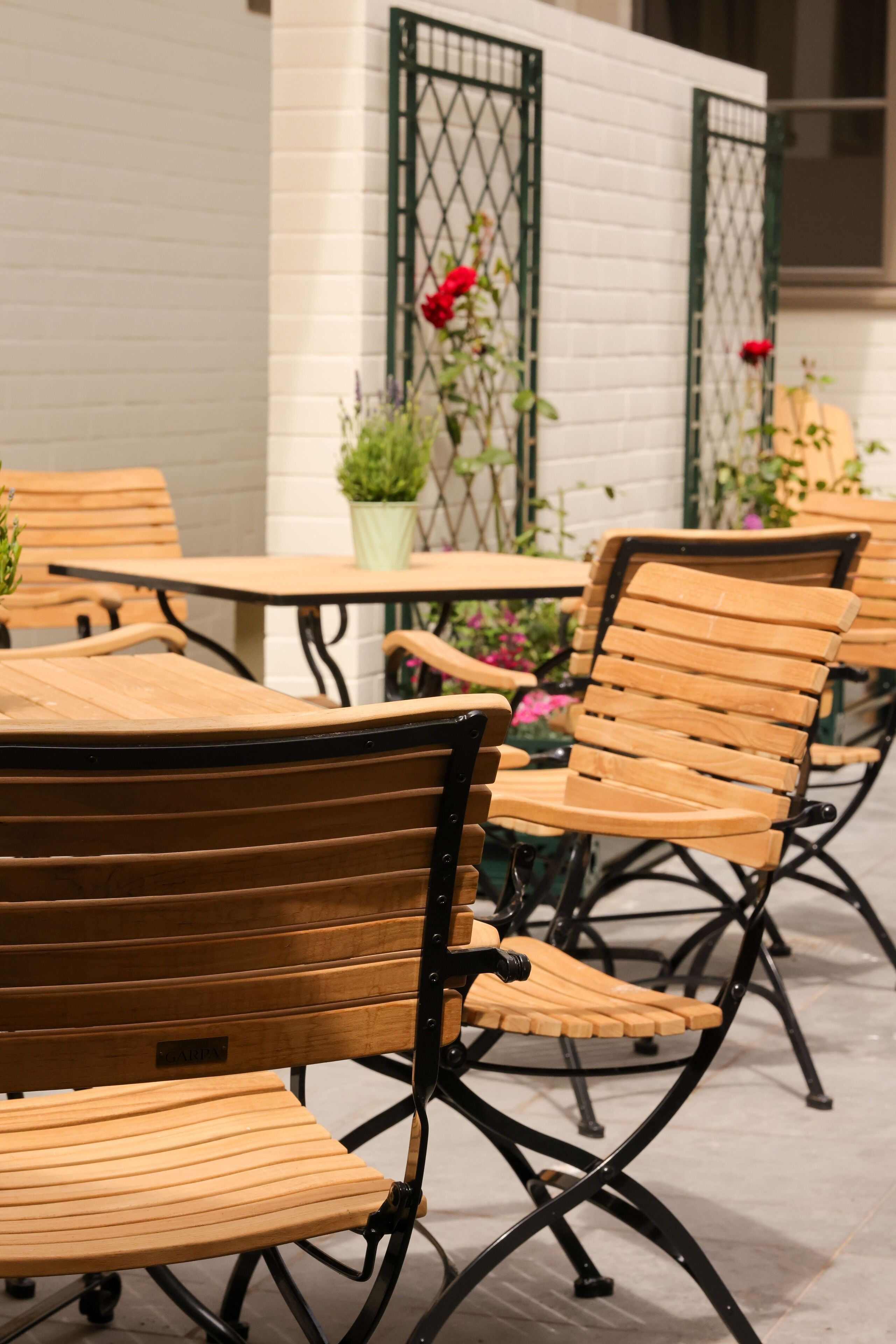 Outdoor wooden chairs and tables with metal frames, arranged in a patio area with a white brick wall and green metal gates.