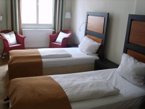 Een hotelkamer met twee eenpersoonsbedden, een nachtkastje, een rode stoel en een raam met gordijnen.