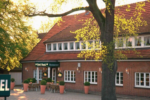Ein Backsteinhaus mit einem Schild, auf dem 'Hotel Maack Restaurant' steht. Vor dem Gebäude befinden sich Tische und Stühle im Freien sowie ein großer Baum.