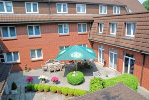 Ein Innenhof mit einem grünen Sonnenschirm mit der Aufschrift 'JEVER', umgeben von Blumentöpfen und einem kleinen Brunnen.