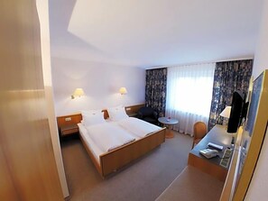 A hotel room with a bed, bedside tables, a desk with a chair, a lamp, and a window with curtains.