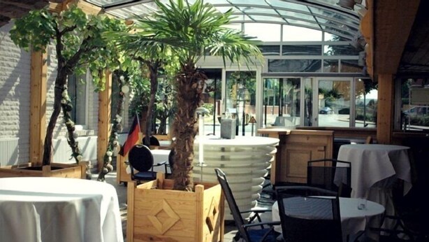 An indoor dining area with tables and chairs, a large central plant, and a glass ceiling. - Hotel & Restaurant Bei Baki (Sehnde)